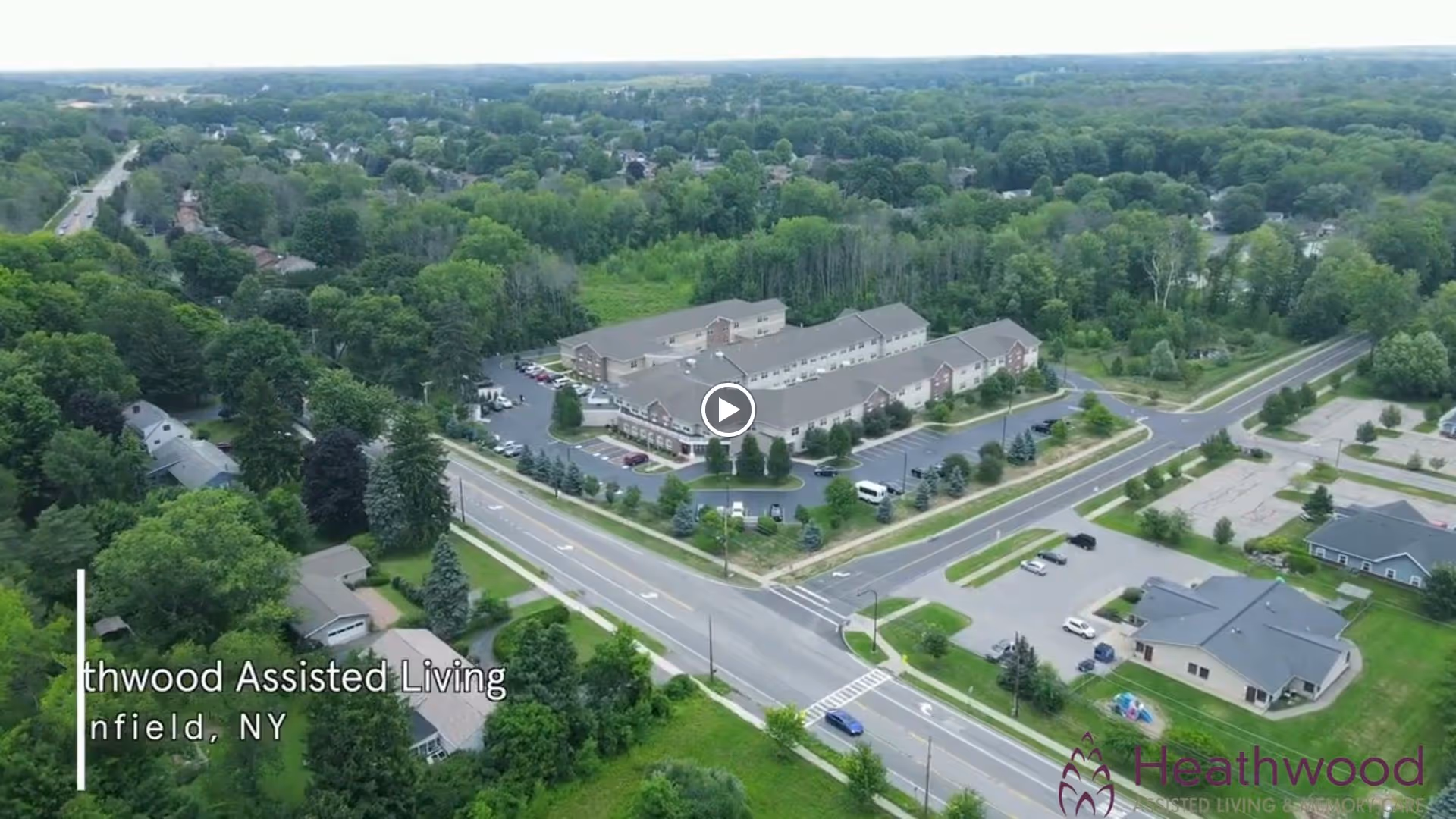 Aerial view of Heathwood Assisted Living and Memory Care facility surrounded by trees and roads in a suburban area of Enfield, NY. The building is large with multiple wings and parking lots, and there is dense greenery and residential houses nearby.