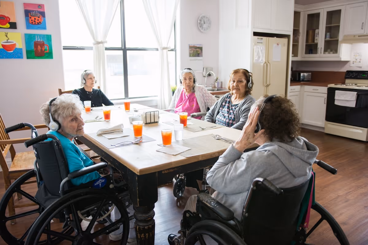 Four elderly women sitting around a wooden dining table in a bright room with large windows. Three of the women are in wheelchairs and all are wearing headphones. The table has placemats, glasses of orange juice, and utensils. The background shows a kitchen area with white cabinets, a refrigerator, and a stove.
