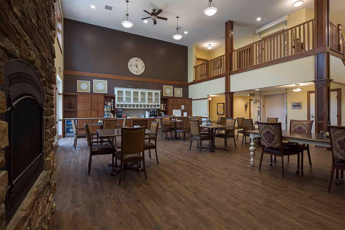 Spacious dining area with multiple wooden tables and chairs, a stone fireplace on the left, a high ceiling with ceiling fans and pendant lights, and a kitchen area with cabinets and a clock on the far wall. There is a second-floor balcony overlooking the dining space.