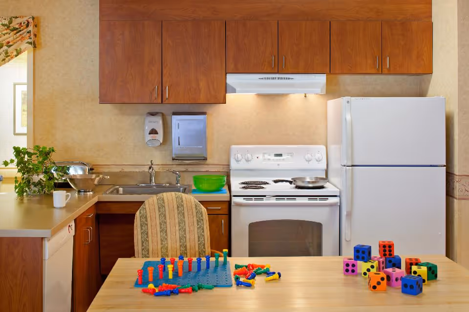 A kitchen area with wooden cabinets, a white electric stove, a white refrigerator, and a sink. On the countertop, there is a green bowl, a metal bowl, a coffee mug, and a small plant. In the foreground, there is a table with colorful pegboard game pieces and large foam dice. A striped upholstered chair is positioned at the table.