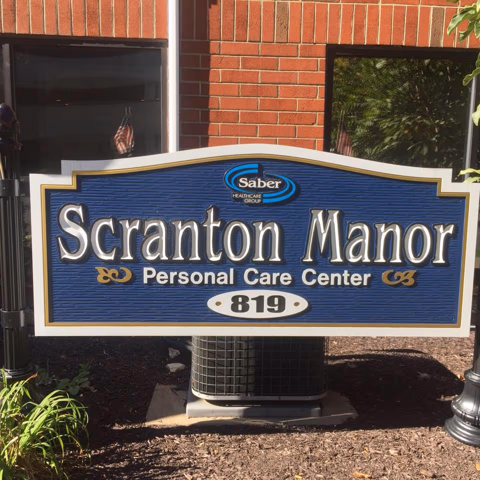 A blue and white sign in front of a brick building that reads 'Scranton Manor Personal Care Center 819' with the Saber Healthcare Group logo above the text.