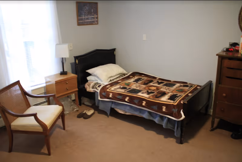 A small bedroom with a single black bed covered with a patterned blanket and pillows. Next to the bed is a wooden nightstand with a lamp and a pair of slippers on the floor. There is a wooden chair with a cushioned seat near a window with white curtains. On the right side, there is a wooden dresser with a digital clock showing 1:25 and some items on top. The walls are light-colored and the floor is carpeted.