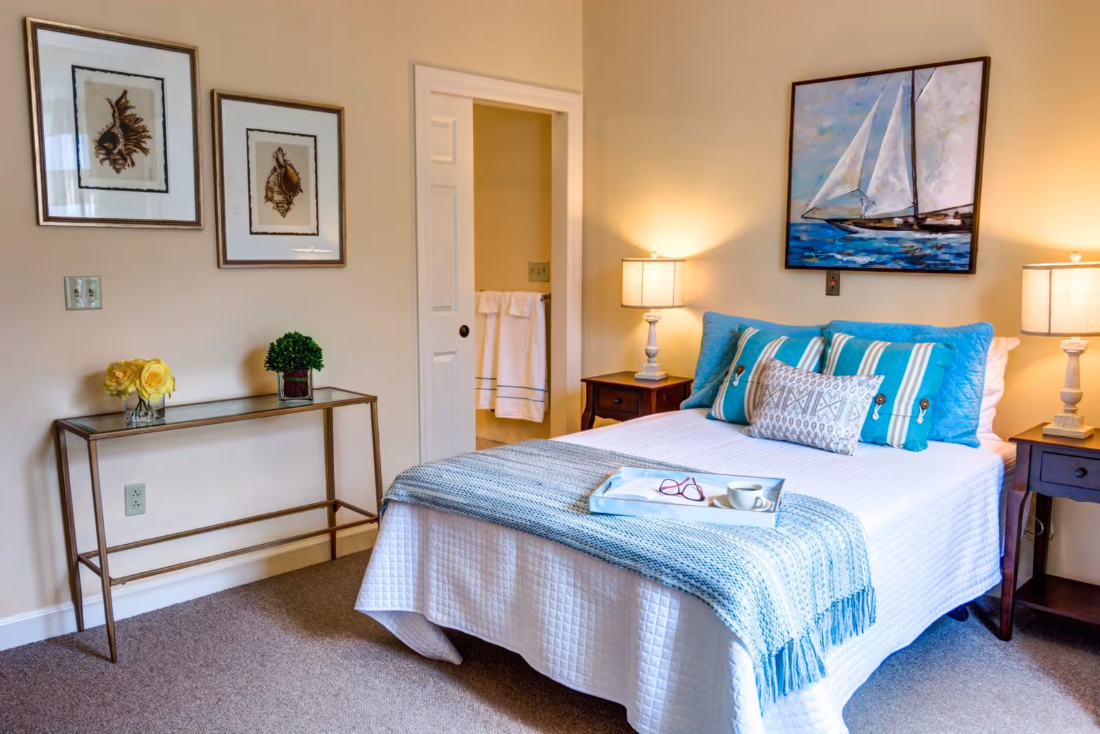 Neatly decorated bedroom with a made bed, striped and patterned pillows, bedside tables with lamps, wall art, and a console table with flowers.