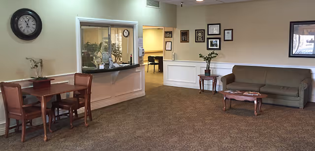 A waiting area with a brown carpeted floor, a small wooden table with two chairs on the left, a reception window with a counter in the center, and a brown sofa with a wooden coffee table on the right. The walls are light-colored with framed certificates and a clock above the table.