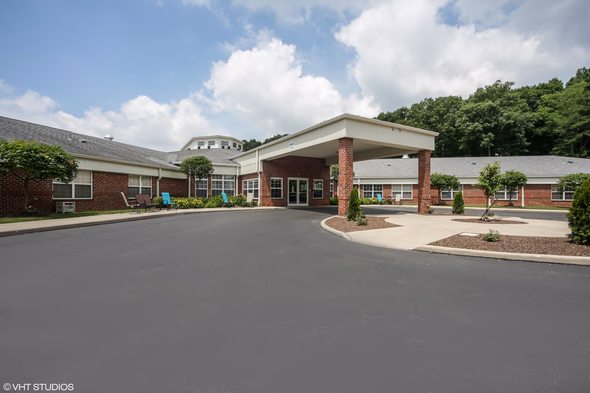Front exterior view of a single-story brick building with a covered entrance, surrounded by a paved driveway and landscaped areas with small trees and shrubs under a partly cloudy sky.