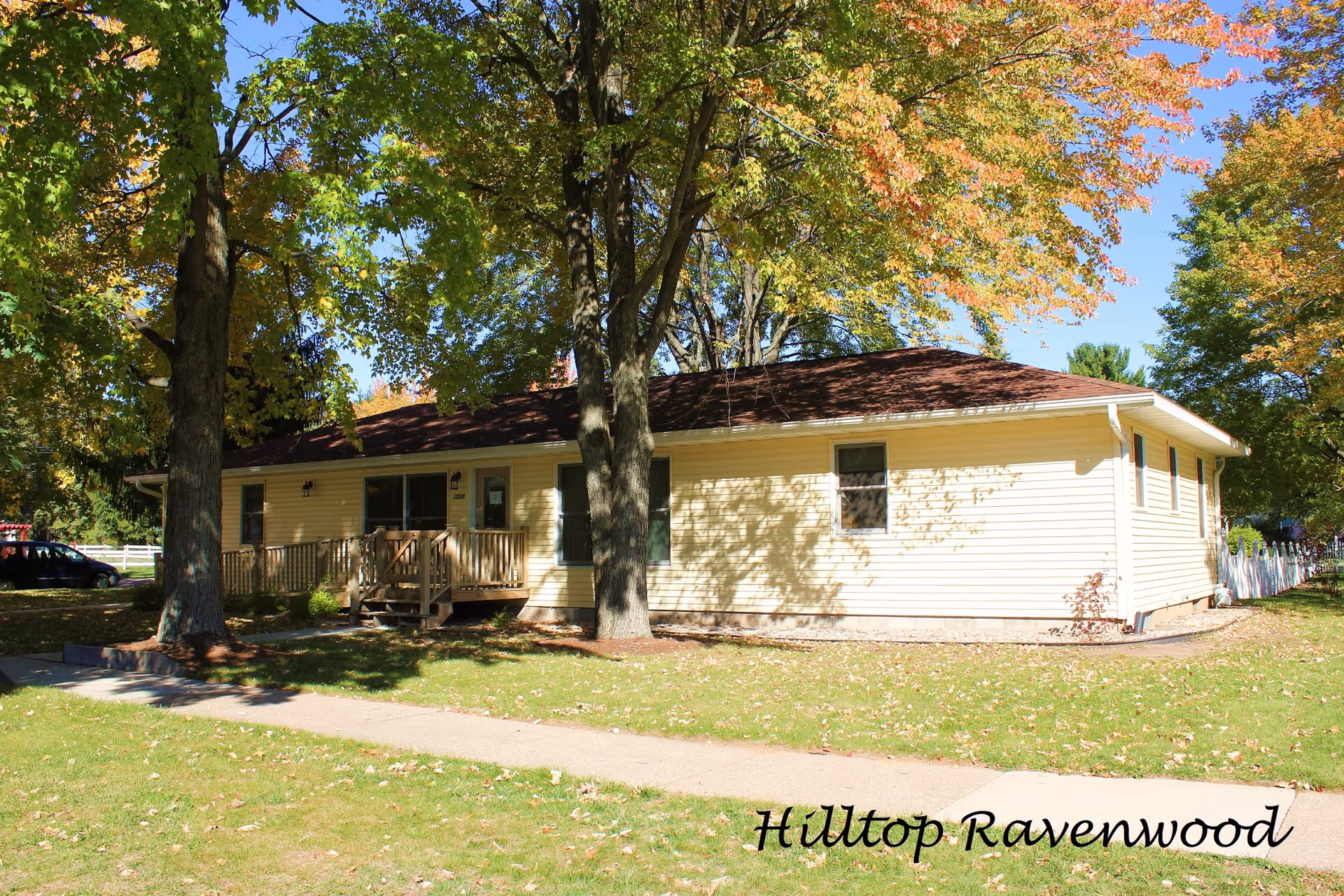 Single-story yellow building with a brown roof surrounded by green grass and large trees with autumn foliage under a clear blue sky. A sidewalk runs in front of the building, and there is a small wooden ramp and steps leading to the entrance.