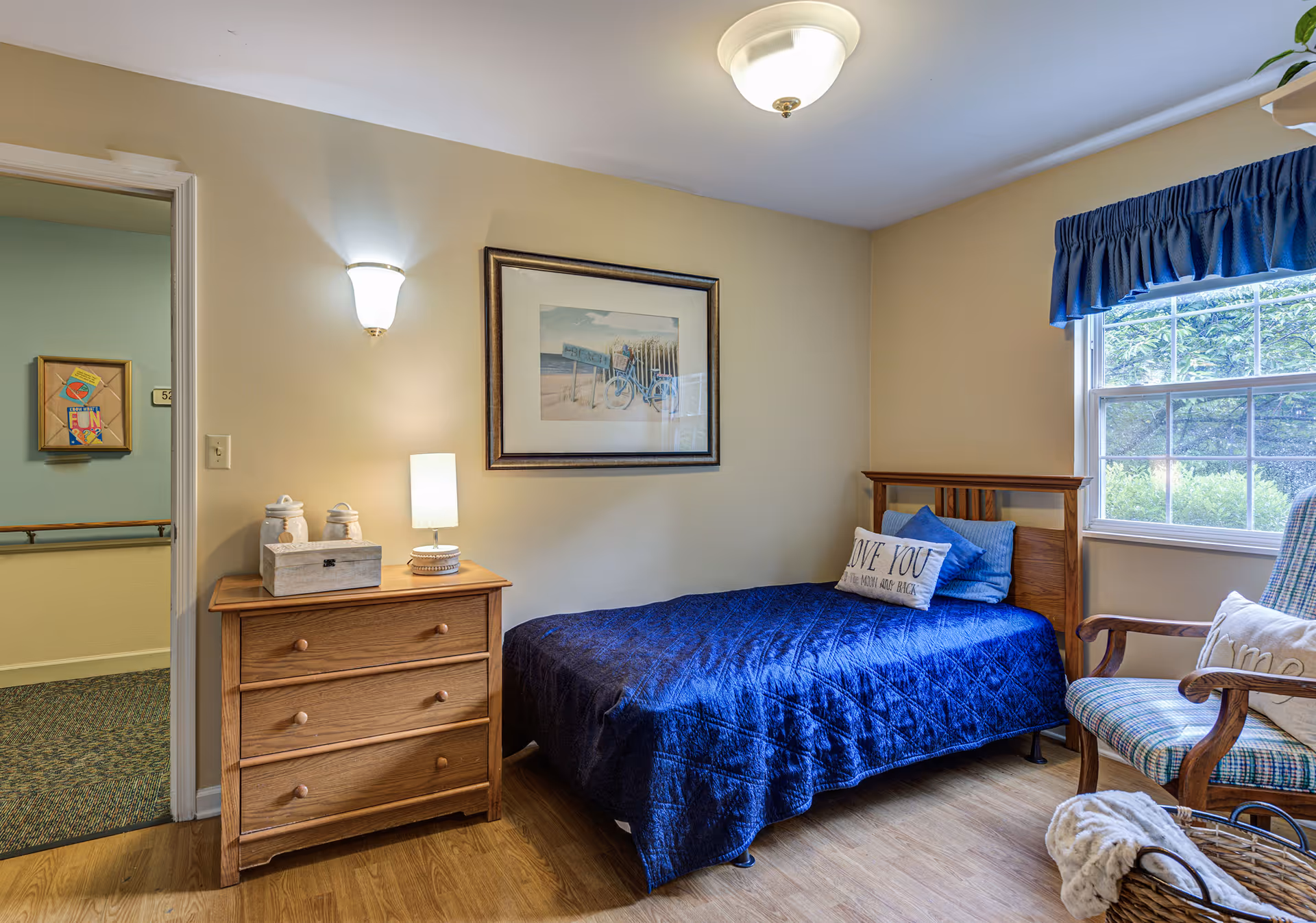 A cozy bedroom in a senior living facility featuring a single bed with a blue quilt and decorative pillows, a wooden dresser with a lamp and decorative jars, a framed beach-themed picture on the wall, a window with blue valance curtains, and a cushioned armchair with a pillow. The room has light yellow walls and wood flooring.