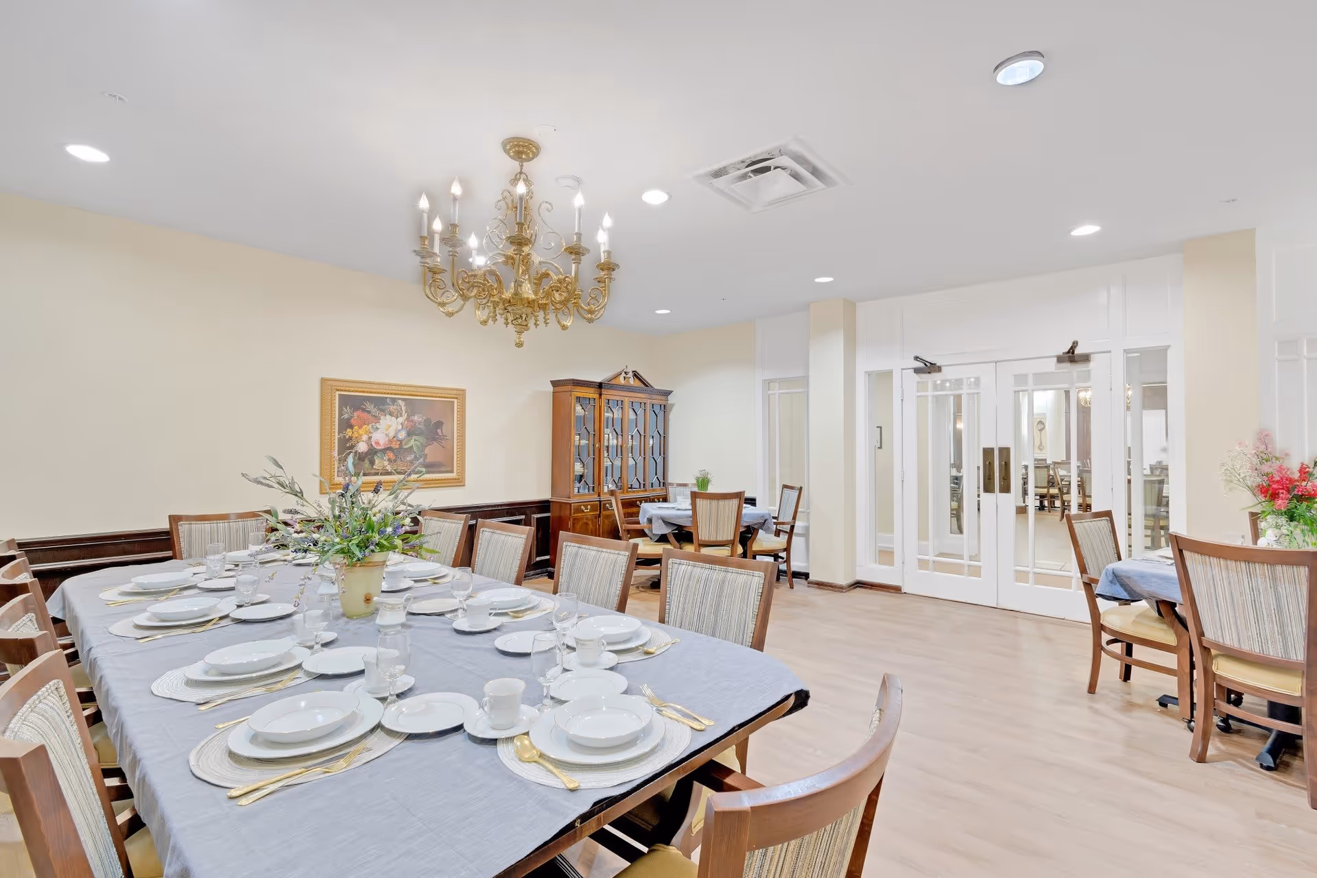 A spacious dining room with a large table set for a meal, featuring white plates, cups, and gold utensils on a gray tablecloth. The room has wooden chairs with striped cushions, a chandelier hanging from the ceiling, a floral painting on the wall, and a wooden china cabinet. There are additional smaller tables with chairs in the background and double glass doors leading to another room.