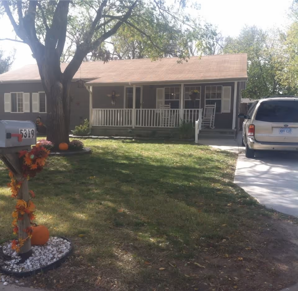 A single-story house with a front porch featuring white railings and rocking chairs. There is a large tree in the front yard, a mailbox decorated with autumn leaves and pumpkins, and a beige SUV parked in the driveway. The lawn is green with some scattered leaves, and the scene is set on a sunny day.
