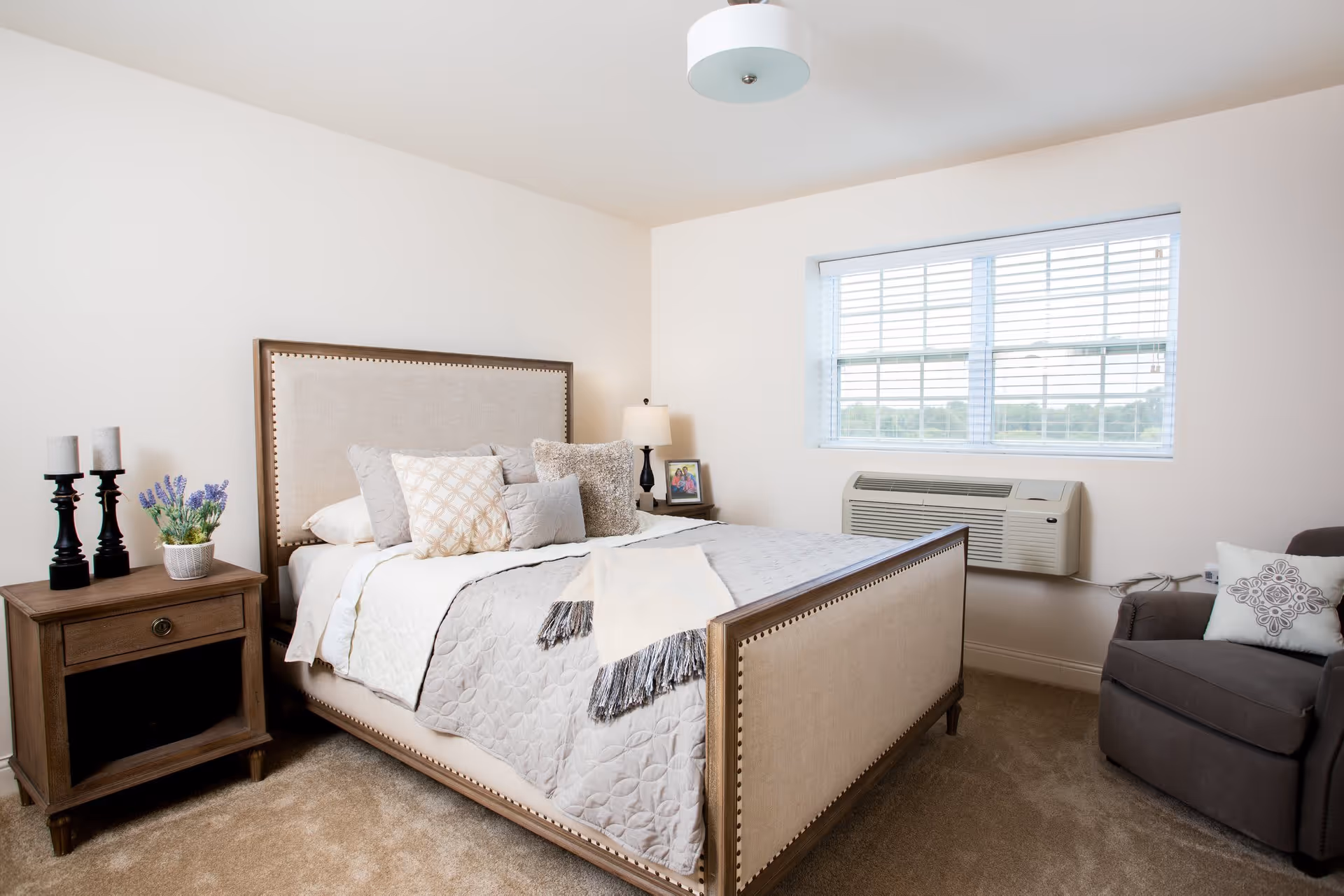 A cozy bedroom with a beige upholstered bed adorned with multiple pillows and a light quilt. Next to the bed is a wooden nightstand with two black candle holders and a small potted plant with purple flowers. A framed photo and a lamp are on the far side of the bed. A window with white blinds lets in natural light, and below it is a wall-mounted air conditioning unit. A gray armchair with a decorative pillow is positioned near the window. The room has beige carpet and white walls.