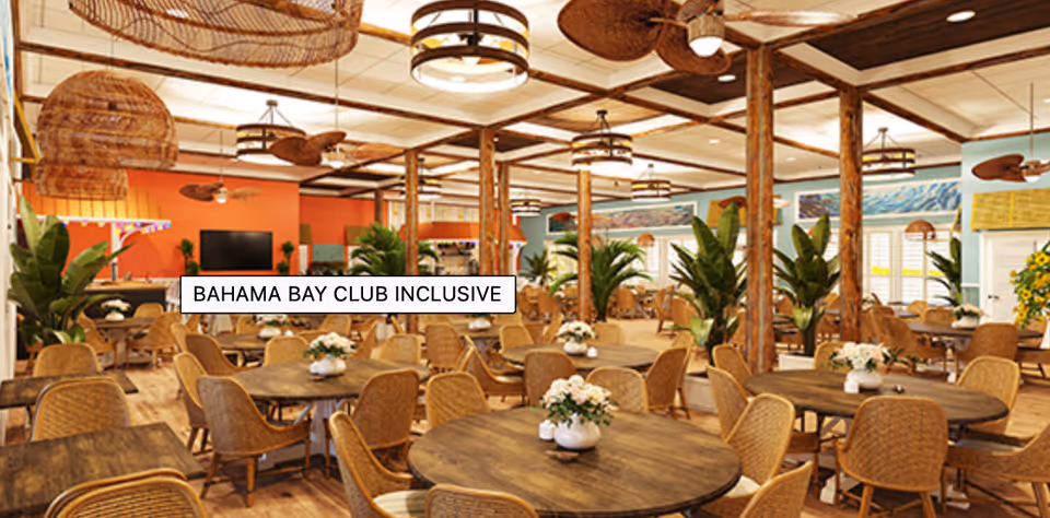 Spacious dining room with multiple round wooden tables and wicker chairs arranged neatly. The room features tropical decor with large green plants, wicker ceiling fans, and warm lighting fixtures. Walls are painted in soft blue and orange tones, creating a welcoming atmosphere.