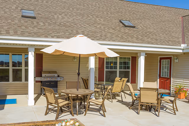 Outdoor patio area with multiple tables and chairs, one table has a large beige umbrella. There is a barbecue grill against the building wall, and the building has beige siding with red shutters and a red door. The sky is clear and blue.