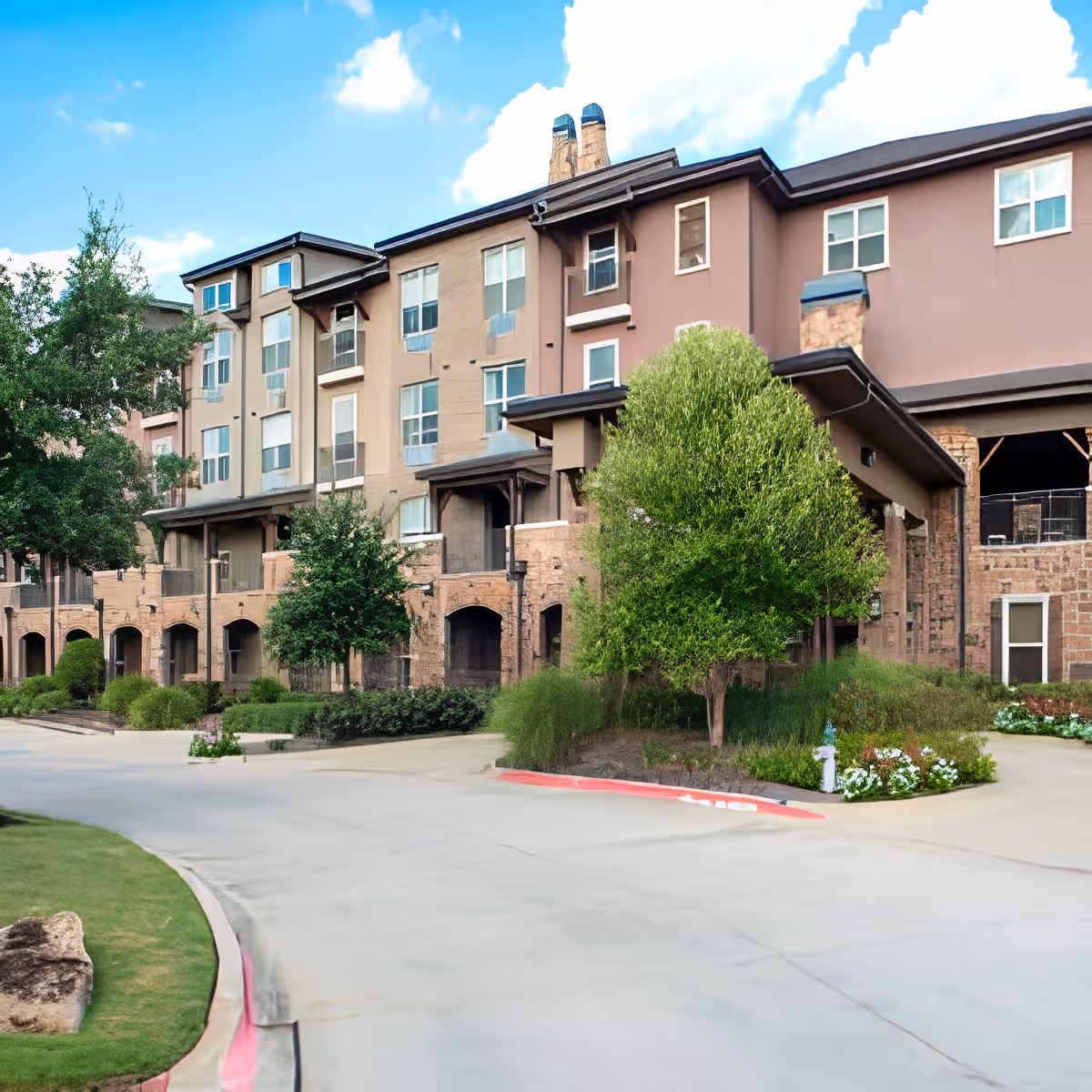 Exterior view of a multi-story senior living facility building with beige and brown walls, multiple windows, and a landscaped area with trees and shrubs in front. The sky is blue with some clouds.