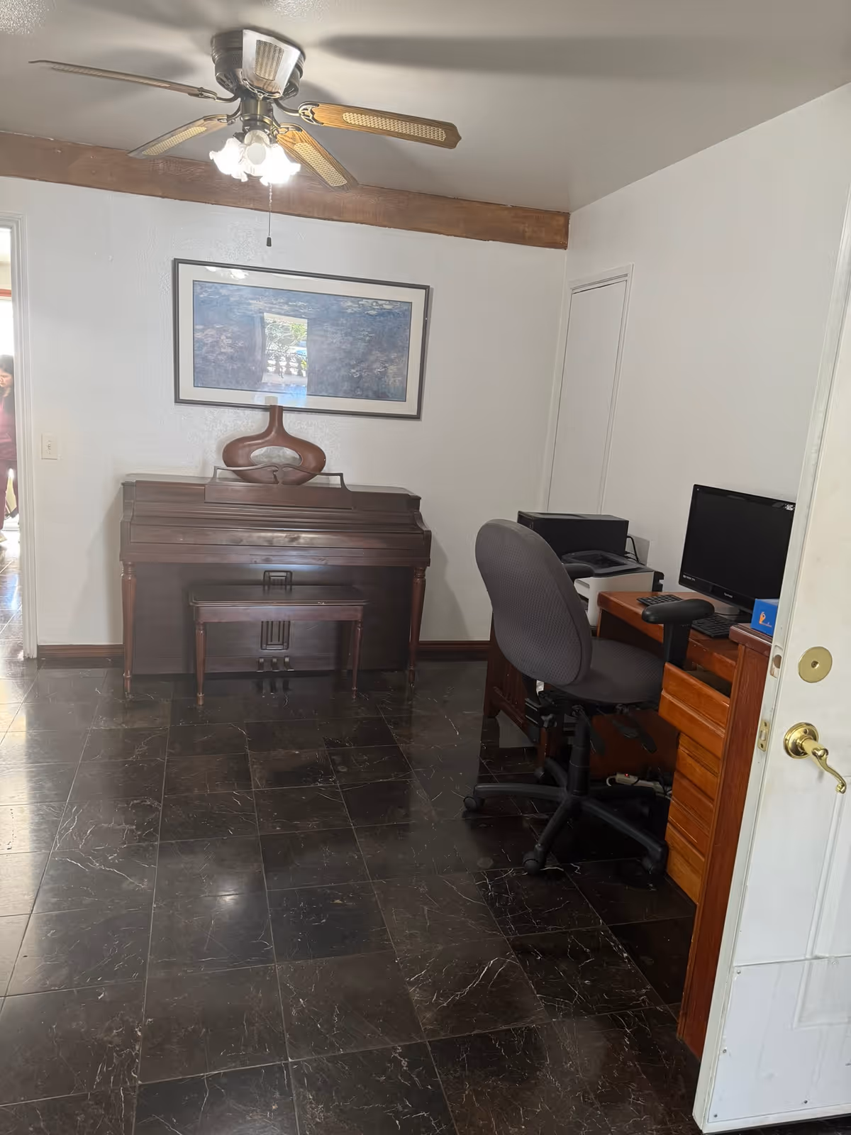 Interior room with an upright piano against the far wall, a desk with a computer and office chair to the right, a ceiling fan overhead, and a glossy dark tiled floor.