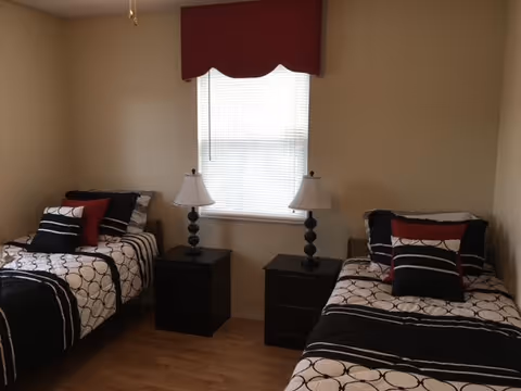 A bedroom with two twin beds separated by two black nightstands, each with a black lamp. The beds have matching black, white, and red bedding. A window with closed blinds and a red valance is centered on the wall behind the nightstands.