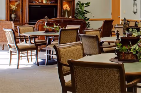 Spacious senior living lounge with round tables, upholstered chairs, and potted plants.