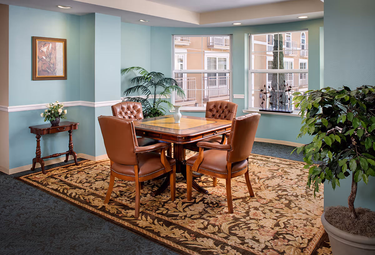 A cozy interior seating area with a wooden square table surrounded by four leather chairs, decorative rug, potted plants and large windows.