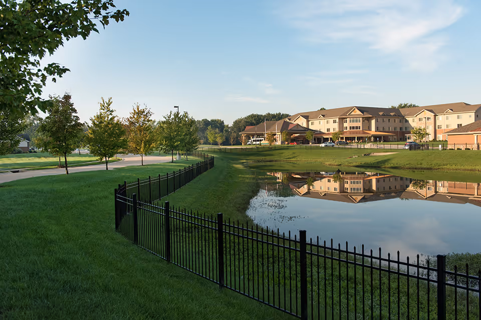 View of a senior living facility named Garden Plaza of Florissant with a large pond reflecting the building, surrounded by green grass, trees, and a black metal fence under a partly cloudy sky.
