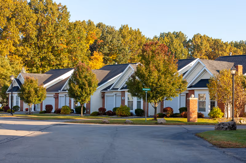 Exterior view of a row of single-story residential buildings with white siding and brick accents, surrounded by trees with autumn foliage and a paved road in the foreground.