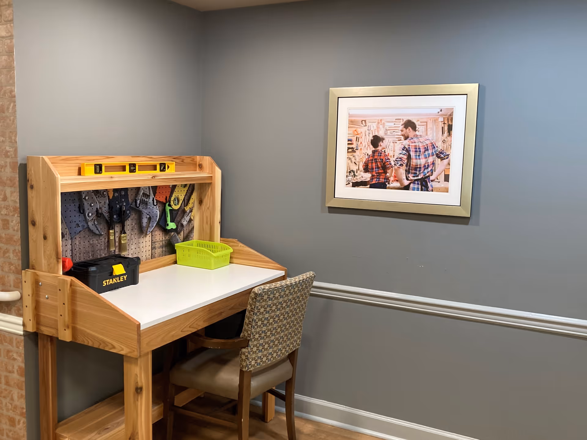 A small wooden workbench with a white surface, a yellow level on top, and various tools hanging on a pegboard behind it. A black Stanley toolbox and a green plastic basket are on the workbench. A patterned chair is placed in front of the workbench. On the gray wall above the chair, there is a framed picture of an adult and a child in a workshop, both wearing plaid shirts.