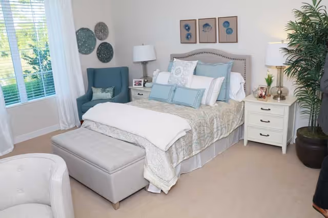Bright, well-decorated bedroom with a made bed, nightstands and lamps, an armchair, ottoman and a large window.