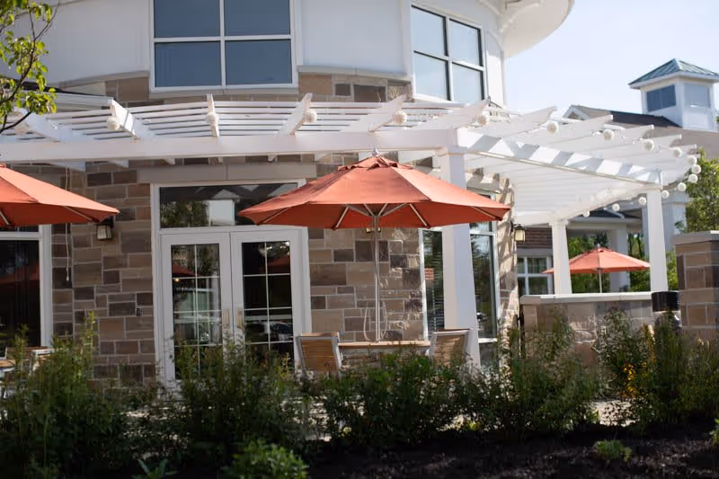 Outdoor patio area at Vista Springs Ravinia Estate featuring stone walls, white pergola, red umbrellas shading wooden tables and chairs, surrounded by greenery and plants.