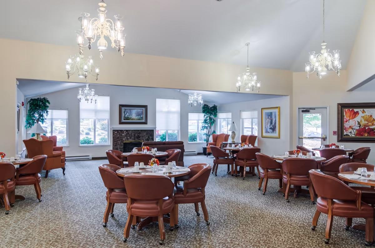 A spacious senior living dining room with multiple round tables surrounded by brown leather chairs. The room features large windows letting in natural light, chandeliers hanging from the ceiling, framed artwork on the walls, and a fireplace in the background. There are also some armchairs and potted plants near the windows.