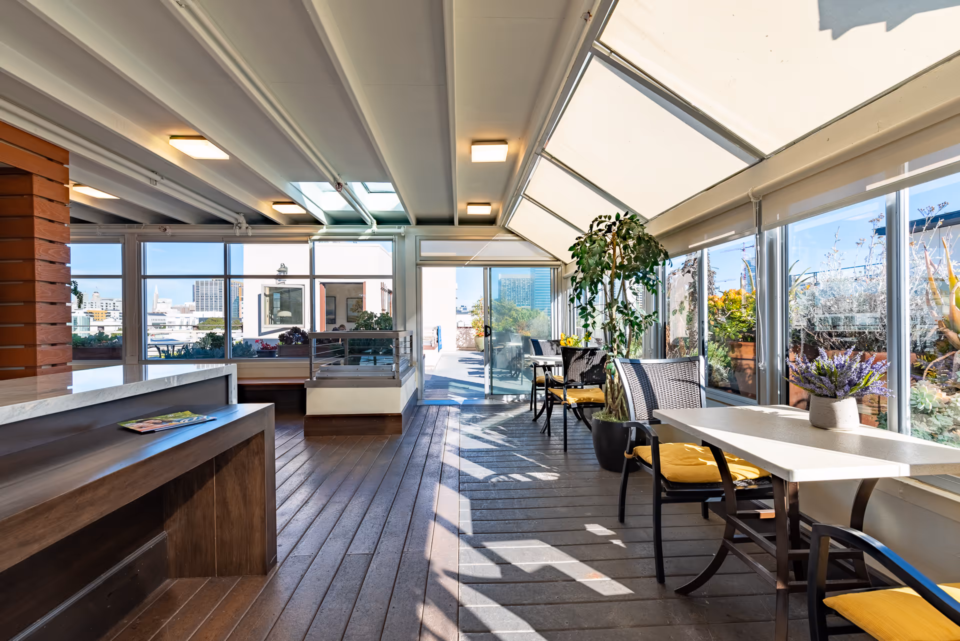 Sunlit enclosed terrace-style dining area with tables, chairs, potted plants and large windows showing city views.