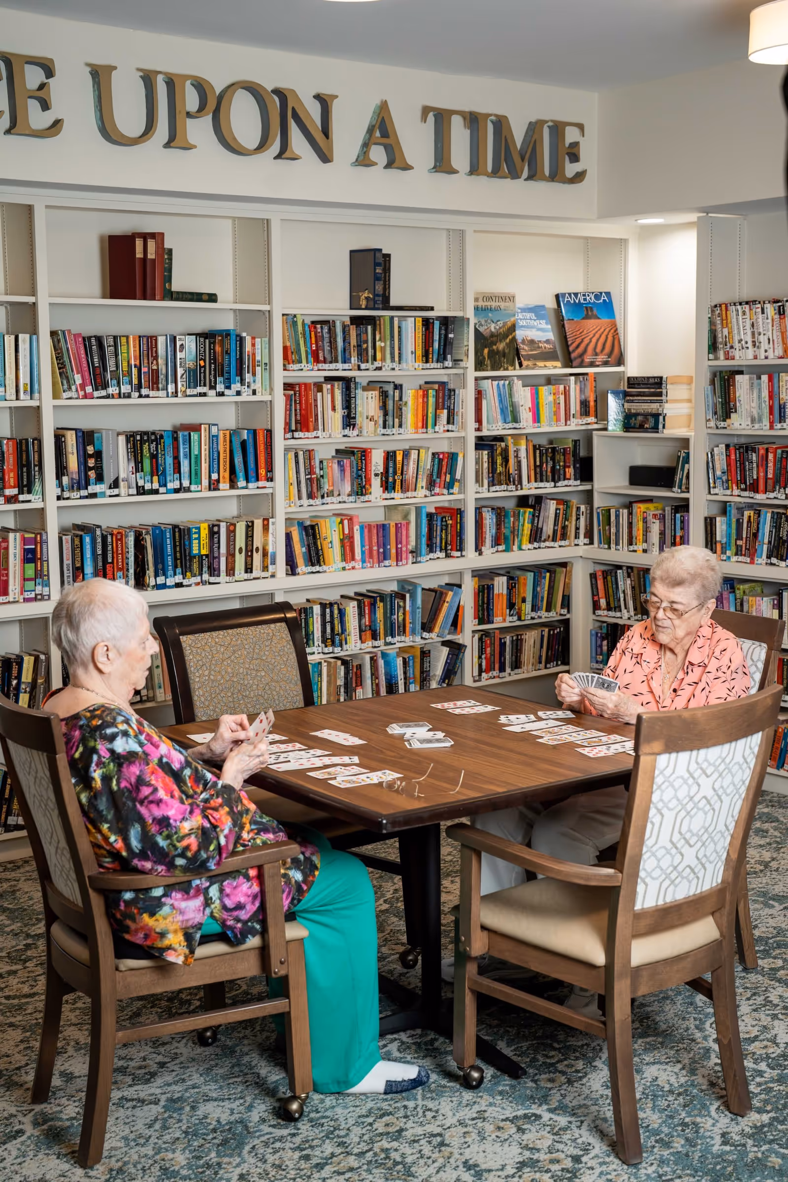 Two elderly women sitting at a wooden table playing cards in a library room with bookshelves filled with books behind them. Above the bookshelves, large letters spell out 'ONCE UPON A TIME'.