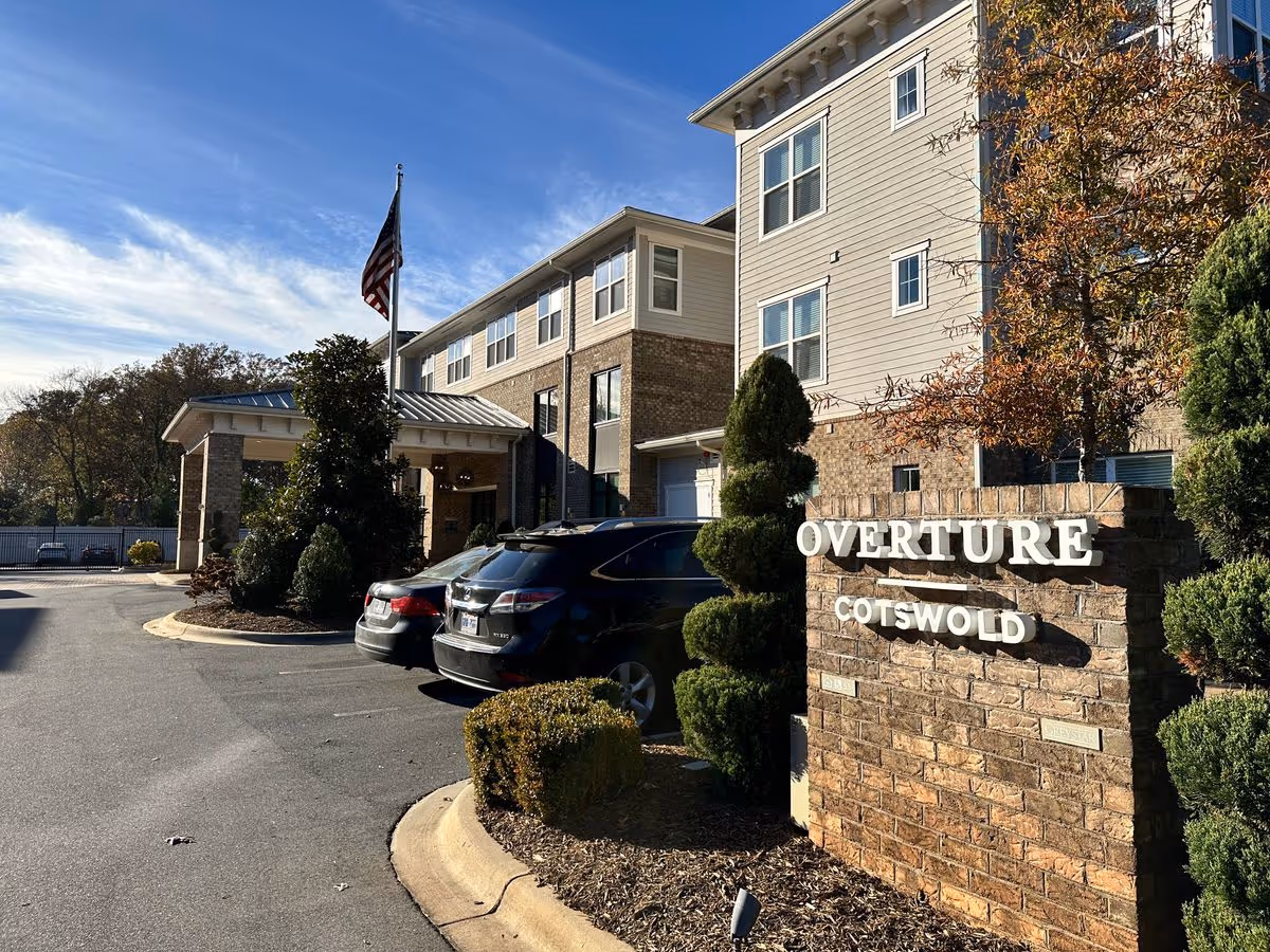Exterior front entrance of the Overture Cotswold building with a brick sign, parked cars, and an American flag.