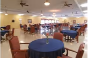 A spacious dining room with multiple round tables covered with blue tablecloths and surrounded by red cushioned chairs. The room has beige walls, ceiling fans, and several light fixtures. Windows with curtains allow natural light to enter the space.