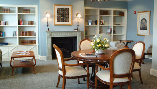 A cozy living room area with a round wooden table surrounded by four upholstered chairs. The table has a flower arrangement and some magazines on it. Behind the table is a fireplace with a framed artwork above it, flanked by two wall sconces. On either side of the fireplace are built-in white bookshelves filled with books and decorative items. The walls are painted light blue, and there is a small wooden side table with magazines on the left side of the image.