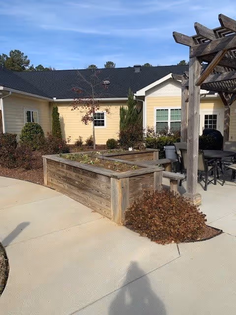 Outdoor patio area with raised wooden garden beds, a pergola, and outdoor seating including a table and chairs. The background shows a yellow building with white trim and windows, surrounded by shrubs and small trees under a clear blue sky.