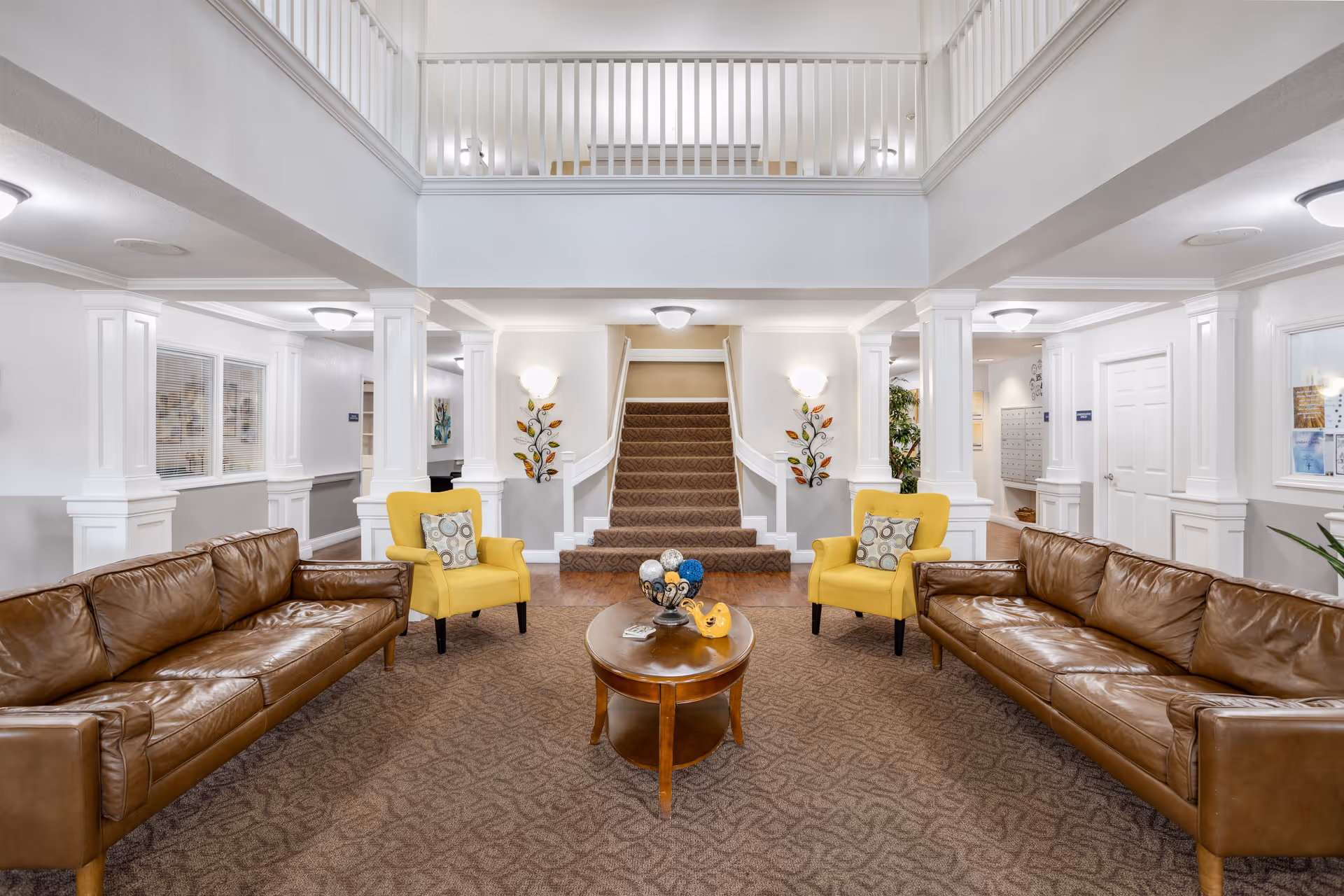 A spacious and well-lit common area with two brown leather sofas and two yellow armchairs arranged around a wooden coffee table. The room features white walls, decorative columns, wall-mounted lights, and a carpeted floor. A staircase with carpeted steps leads to an upper level with a white railing.