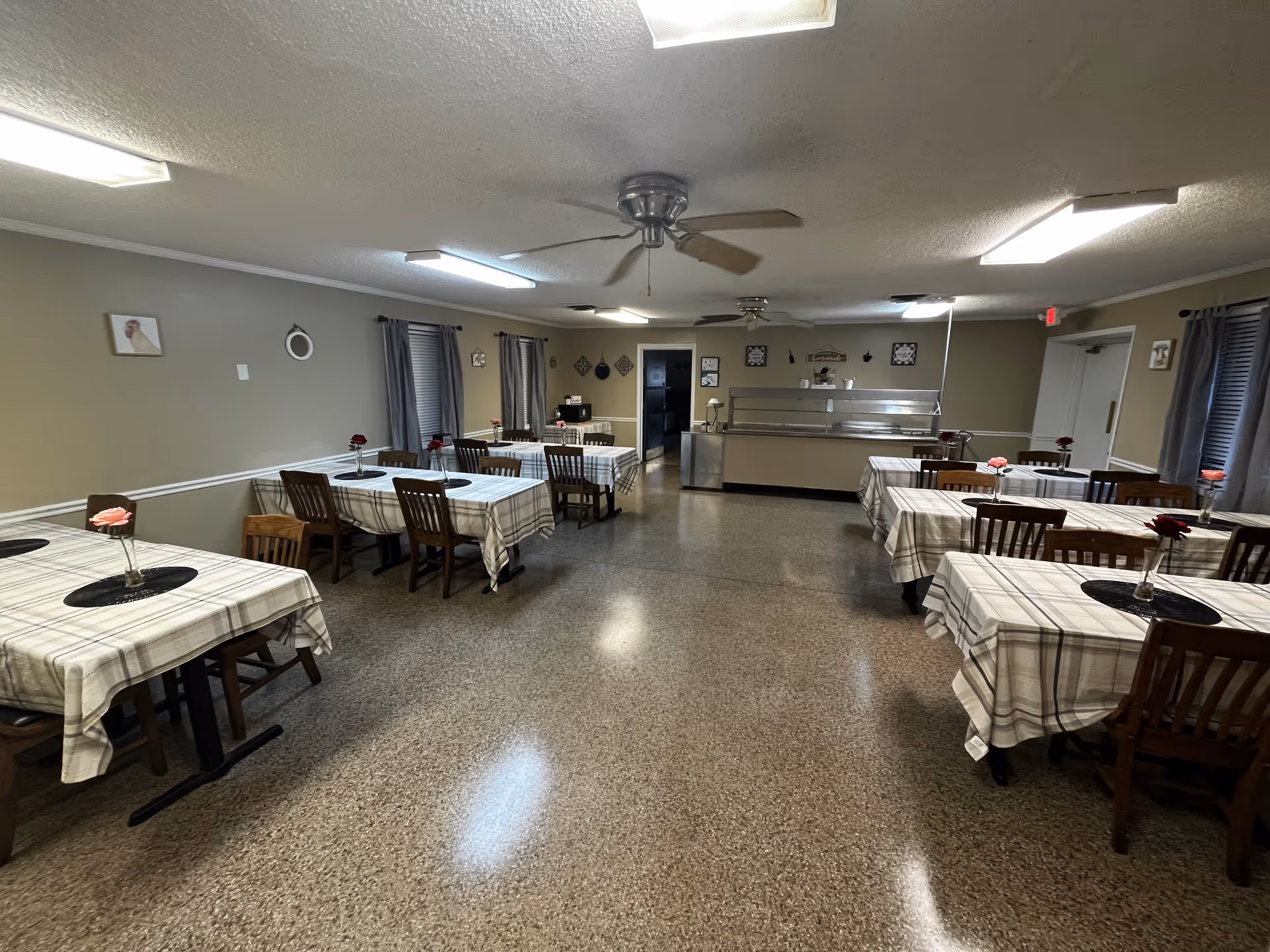 Interior view of a dining room with multiple tables covered in plaid tablecloths, each table having a small vase with a single flower. The room has beige walls, several windows with gray curtains, ceiling fans, and fluorescent lighting. At the far end, there is a serving counter with a metal food shield.