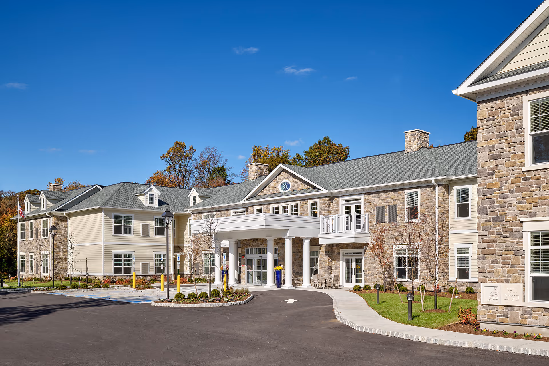 Two-story stone-and-siding senior living building with a covered main entrance, circular driveway and landscaped grounds under a clear blue sky.