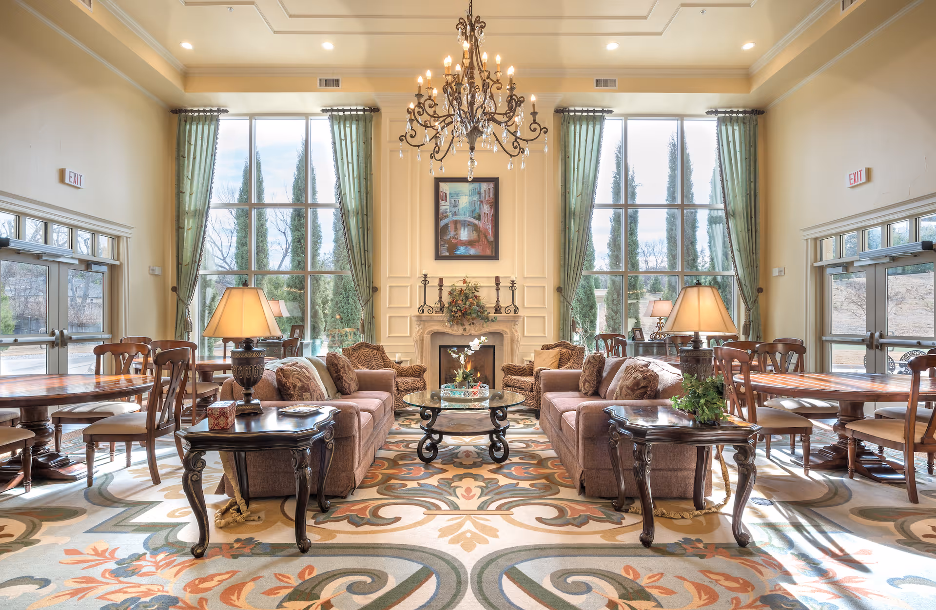 A spacious and elegant living room area with two brown sofas facing each other, a glass coffee table in the center, and two side tables with lamps. The room features large floor-to-ceiling windows with green curtains, a decorative fireplace with floral arrangement and a framed painting above it, and a large ornate chandelier hanging from the ceiling. There are also wooden dining tables and chairs on either side of the room, and exit doors on both sides.