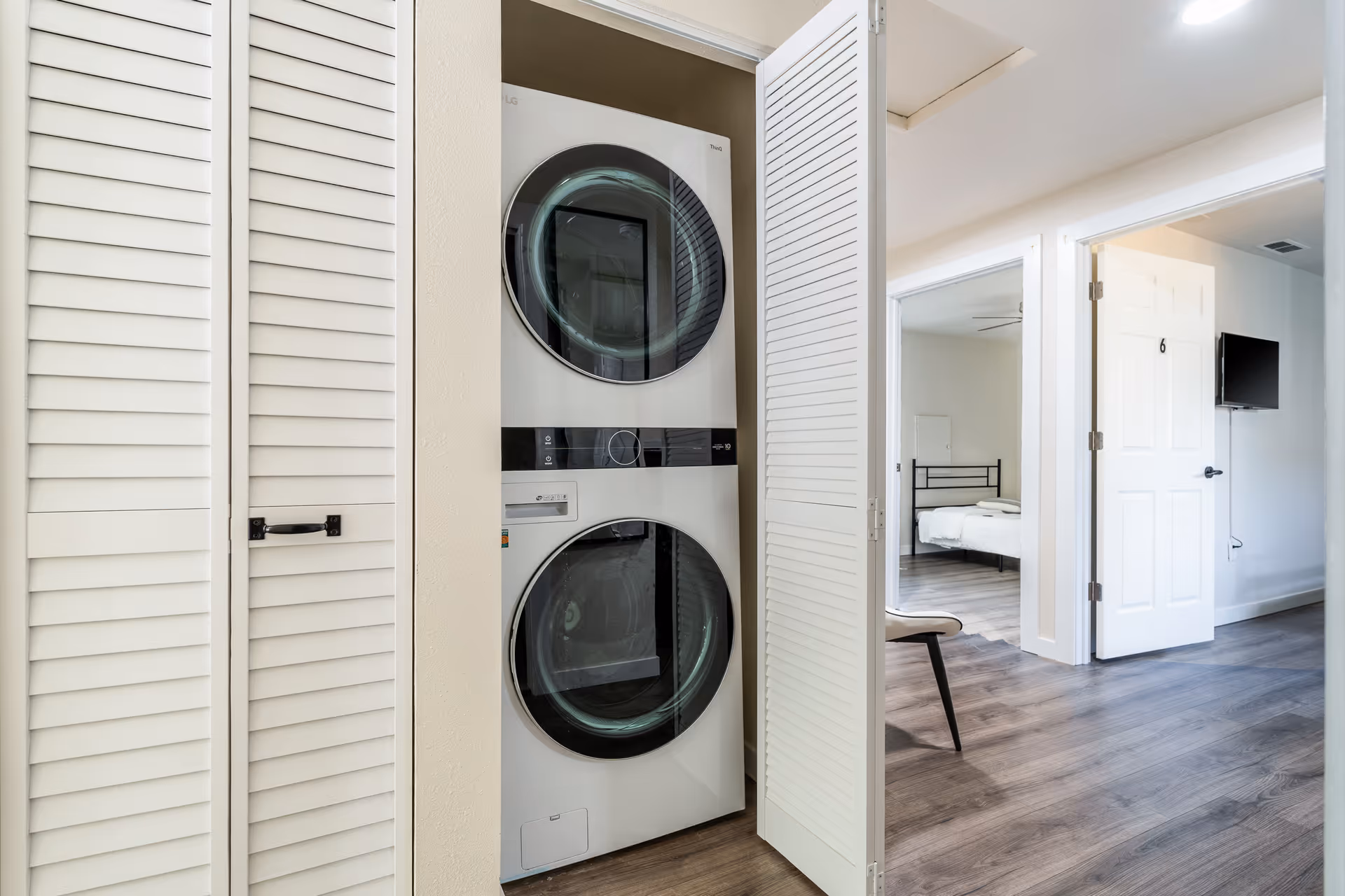 A laundry closet with a stacked washer and dryer behind white louvered doors, adjacent to a hallway leading to a bedroom with a bed and a wall-mounted TV.