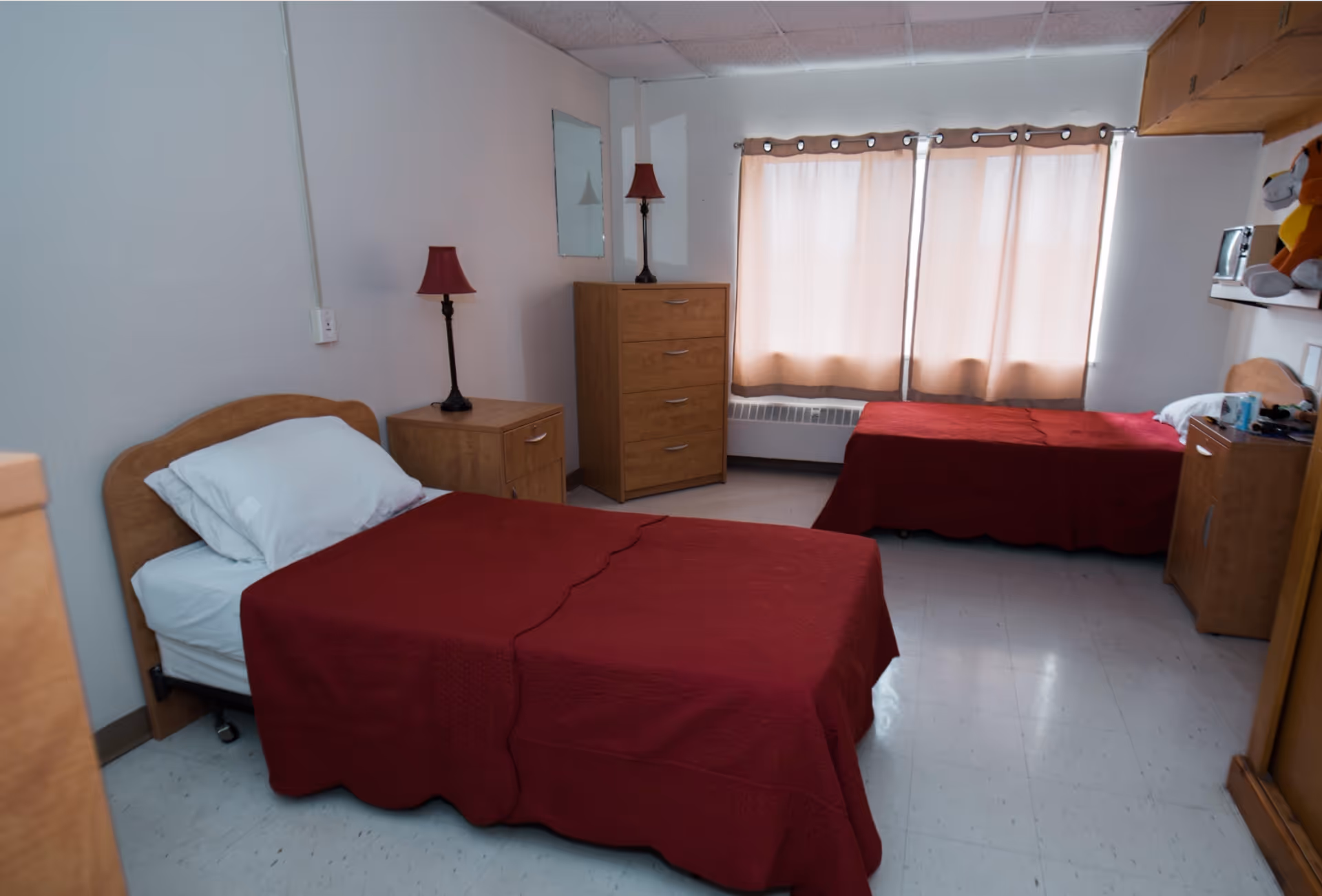 Two twin beds with red bedspreads in a simple furnished bedroom with wooden dressers and a curtained window.
