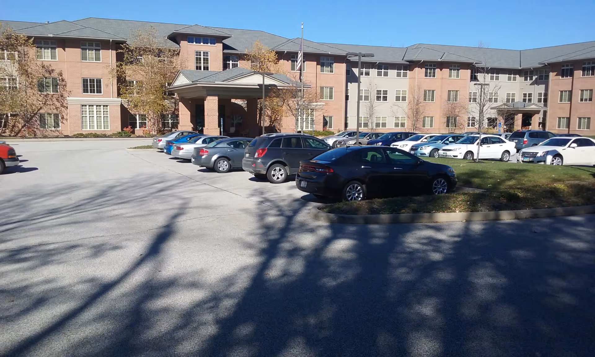 Front exterior of a three-story senior living building with a covered entrance, flagpole, and a parking lot full of cars under a clear blue sky.