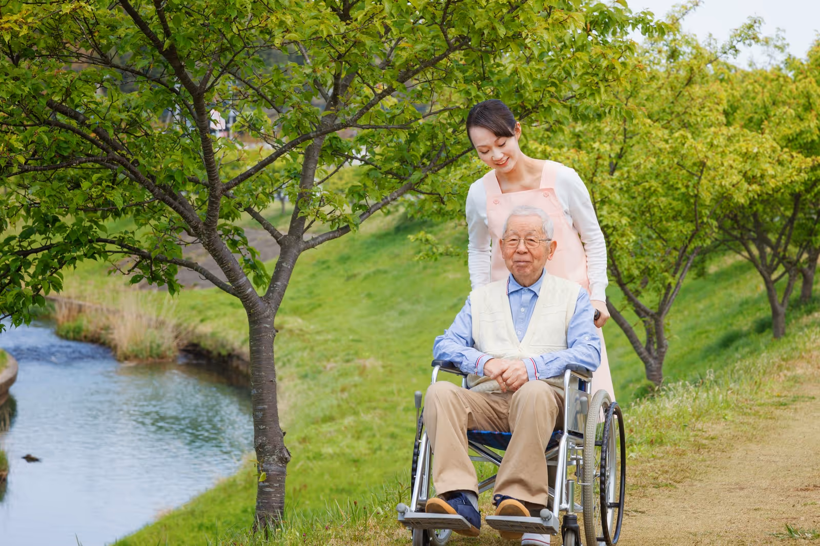 An elderly man sitting in a wheelchair being pushed by a smiling caregiver along a grassy path lined with trees next to a small stream.