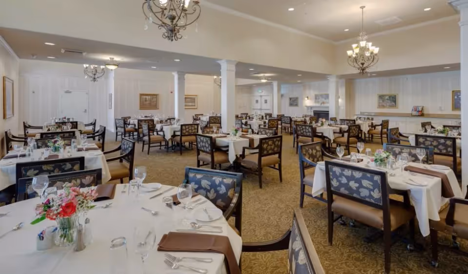 Spacious dining room with multiple tables dressed in white tablecloths, floral centerpieces, chandeliers, and patterned chairs.
