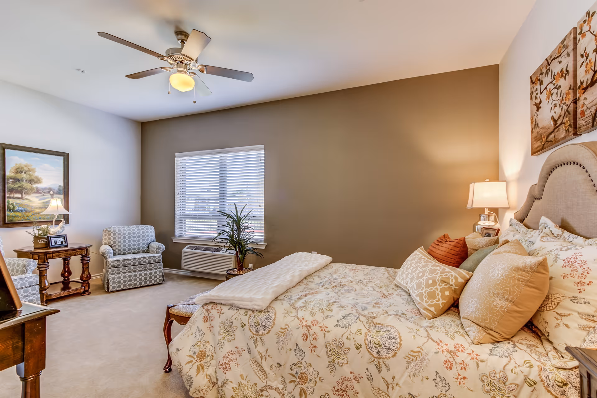 A cozy bedroom in an assisted living facility featuring a bed with floral patterned bedding and multiple pillows, a ceiling fan with light, two patterned armchairs, a wooden side table with a lamp and framed photo, a window with blinds, a potted plant, and wall art above the bed.