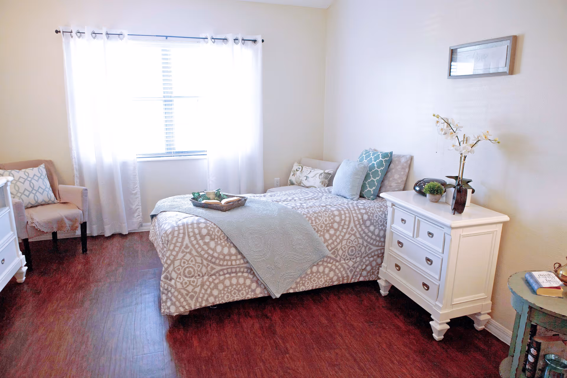 A bright bedroom with a single bed covered in patterned bedding and multiple pillows. Next to the bed is a white nightstand with decorative plants and a vase with flowers. A window with white curtains lets in natural light. There is a chair with a cushion and a small round table with books on the right side of the room. The floor is wooden.