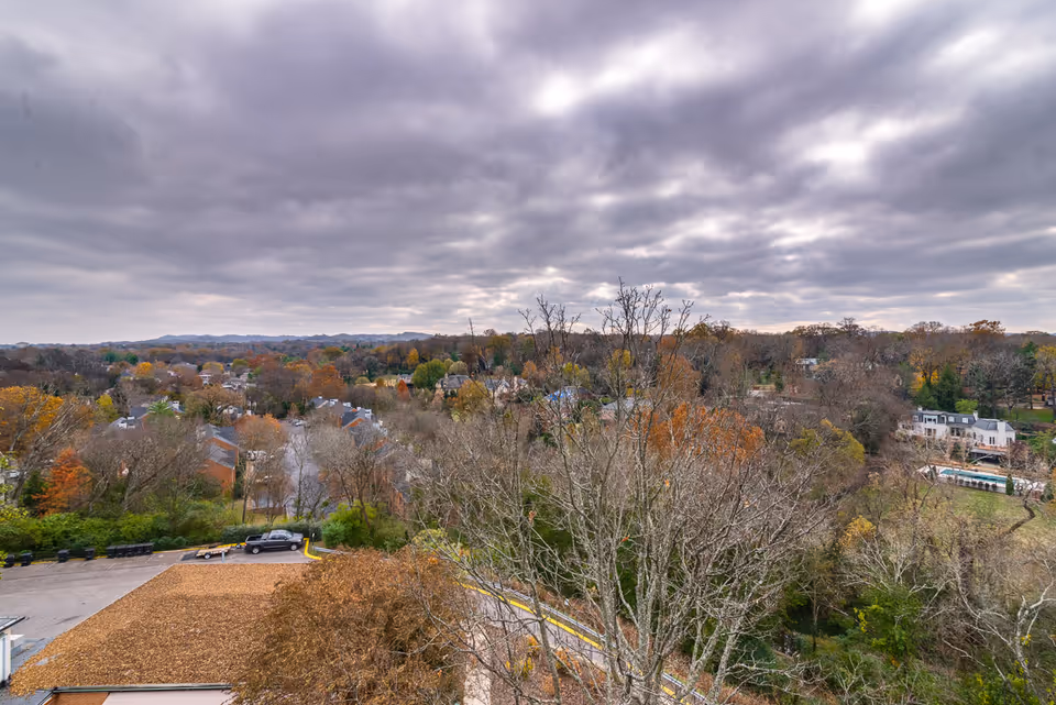 Aerial view of a residential area with many trees showing autumn colors under a cloudy sky. The foreground includes the roof of a building and a parking lot with a few vehicles.