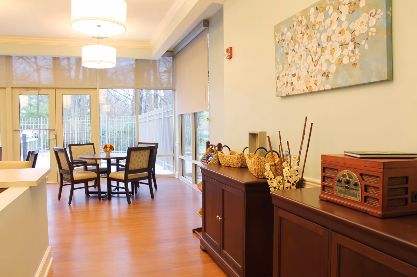 A bright and cozy dining area with a round table surrounded by four cushioned chairs. Large windows with roller shades allow natural light to fill the room. On the right side, there are dark wooden cabinets with decorative baskets, a vintage-style radio, and a floral painting hanging on the wall. The floor is wooden, and two modern pendant lights hang from the ceiling.