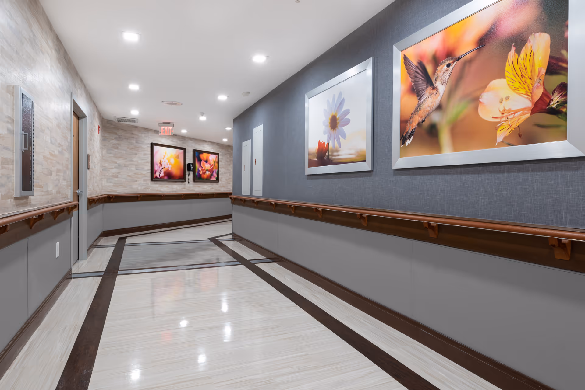 A clean, well-lit hallway in a senior living facility with handrails on both sides and framed floral artwork on the walls. The floor has a polished, light-colored surface with dark border accents. The hallway leads to a corner with two more framed pictures and an exit sign above a door.