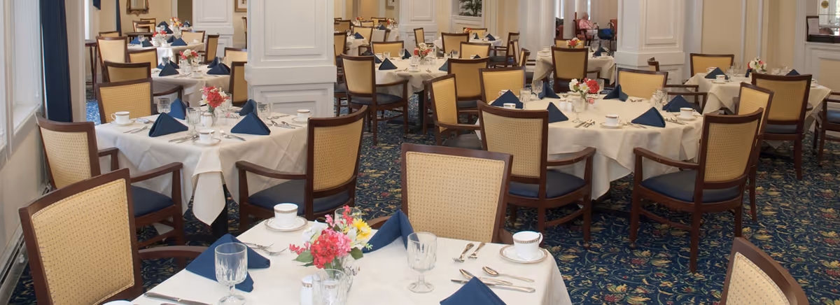 A formal dining room with multiple round tables covered with white tablecloths, each set with blue folded napkins, cups, glasses, silverware, and small floral centerpieces. The room has upholstered wooden chairs and a patterned blue carpet.