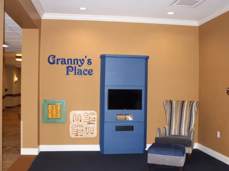 A cozy corner in a senior living facility with a blue cabinet holding a TV, a striped armchair with a matching ottoman, and two wall-mounted activity boards. The wall is painted beige with the words 'Granny's Place' in blue letters.