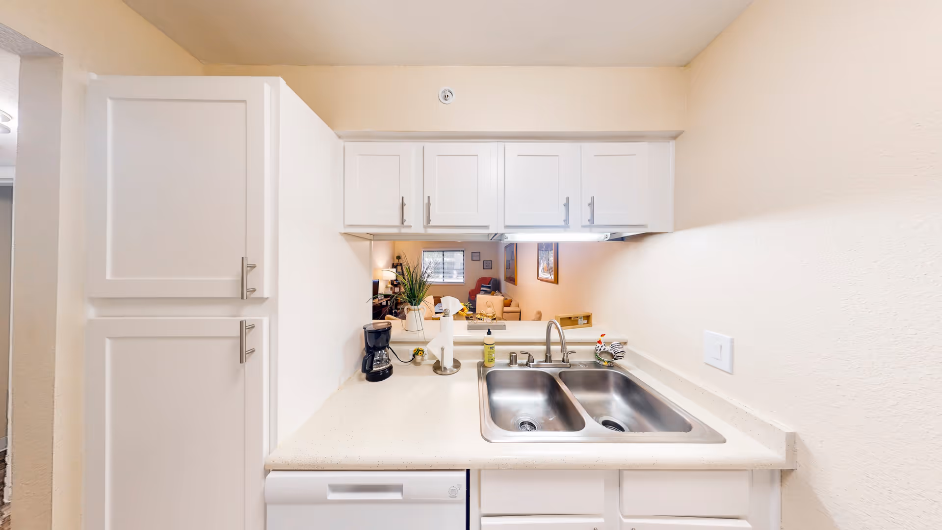 A clean kitchen area with white cabinets and countertops, featuring a double stainless steel sink with a faucet. On the countertop, there is a coffee maker, a paper towel holder, a small plant, and a soap dispenser. Above the sink, there are white upper cabinets and a pass-through window that looks into a living room with furniture and wall art.